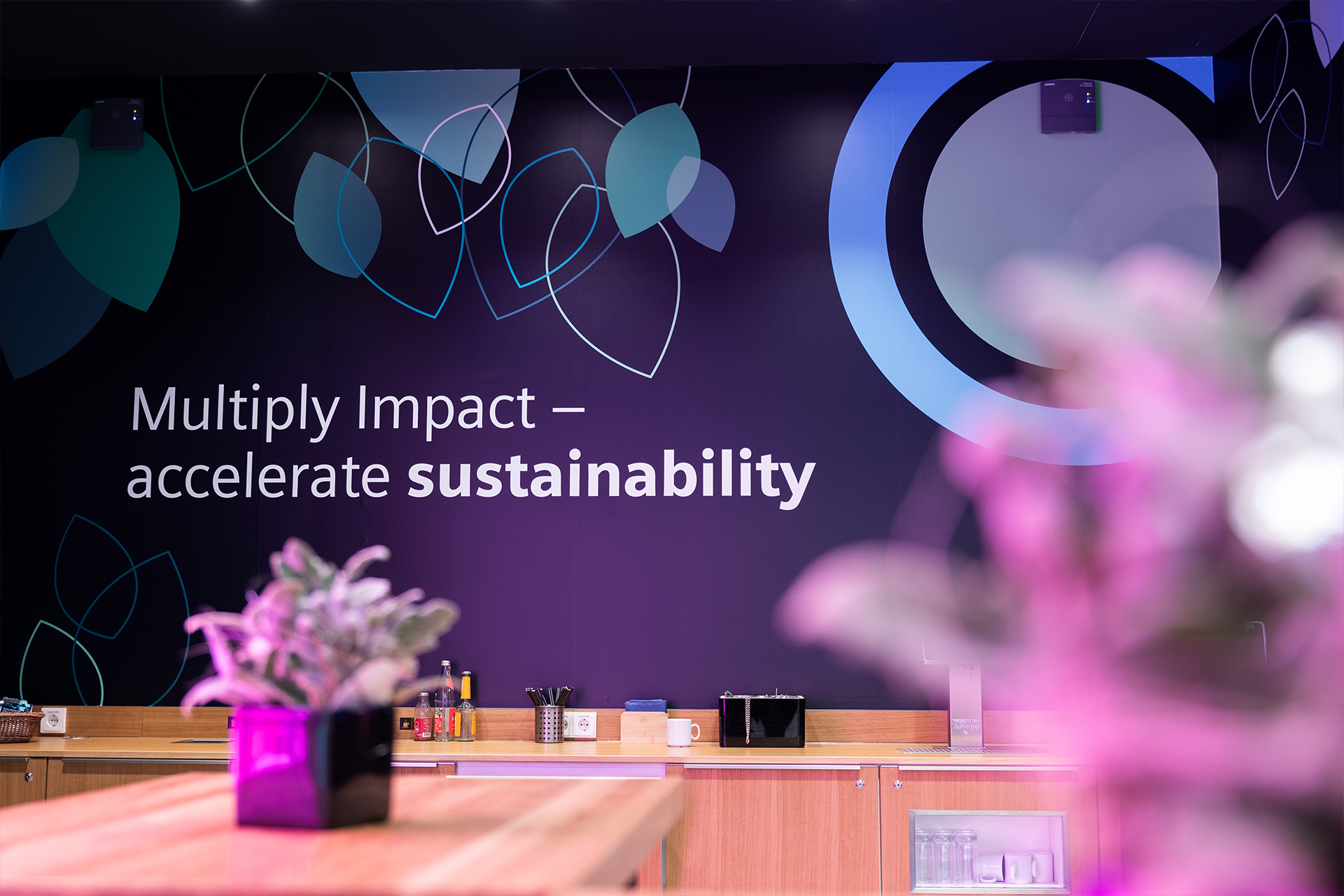 Eine Theke ist zu sehen, im Hintergrund eine große Leinwand mit Schriftzug „Multiply Impact – accelerate sustainability“