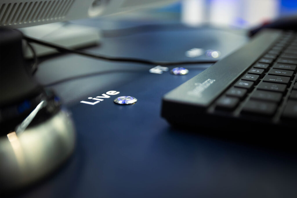 Detailaufnahme eines schwarzen Bedienfelds mit dem Wort „Live“, beleuchteten Knöpfen und einer Tastatur im Vordergrund. Im Hintergrund ist ein Monitorfuß zu erkennen. Die Szene vermittelt einen modernen Arbeitsplatz oder eine interaktive Steuereinheit.