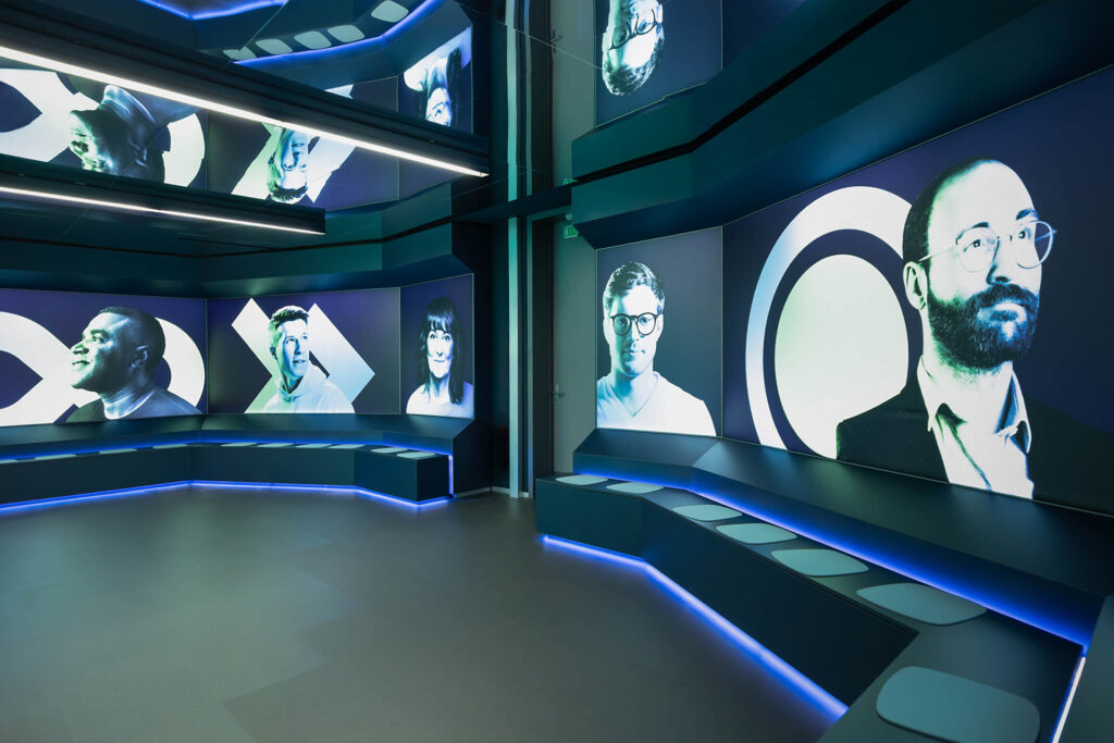 Panoramaaufnahme eines futuristischen Kinosaals mit gebogener Sitzbank und großflächigen LED-Wänden, die stilisierte Porträts von Menschen in blau-grüner Ästhetik zeigen. Die Sitznischen sind beleuchtet, der Raum ist immersiv inszeniert.