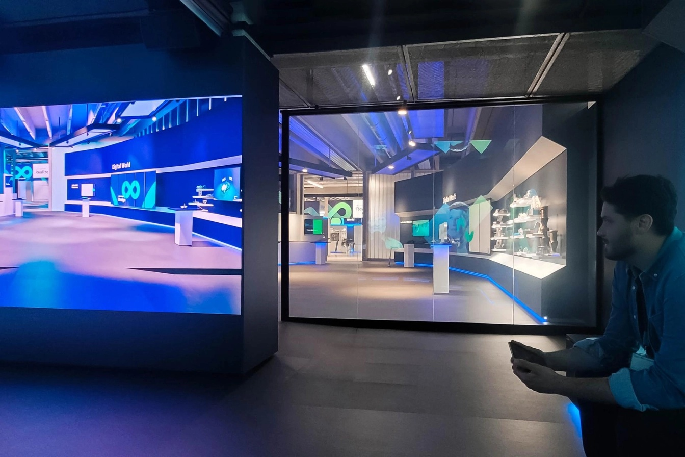 Blick in einen abgedunkelten Raum mit großem Bildschirm, auf dem eine virtuelle Ausstellungsfläche zu sehen ist. Rechts sitzt ein Besucher mit einem Controller in der Hand, der den Raum beobachtet. Die Darstellung vermittelt ein Mixed-Reality-Erlebnis.