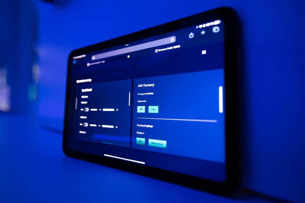 Tablet mit geöffneter Raumsteuerungsoberfläche, auf dem sich unter anderem der Bereich „AM Factory“ aktivieren lässt. Die Benutzeroberfläche zeigt verschiedene Schalter und Regler zur Licht- und Displaysteuerung, eingebettet in eine tiefblaue Umgebung.