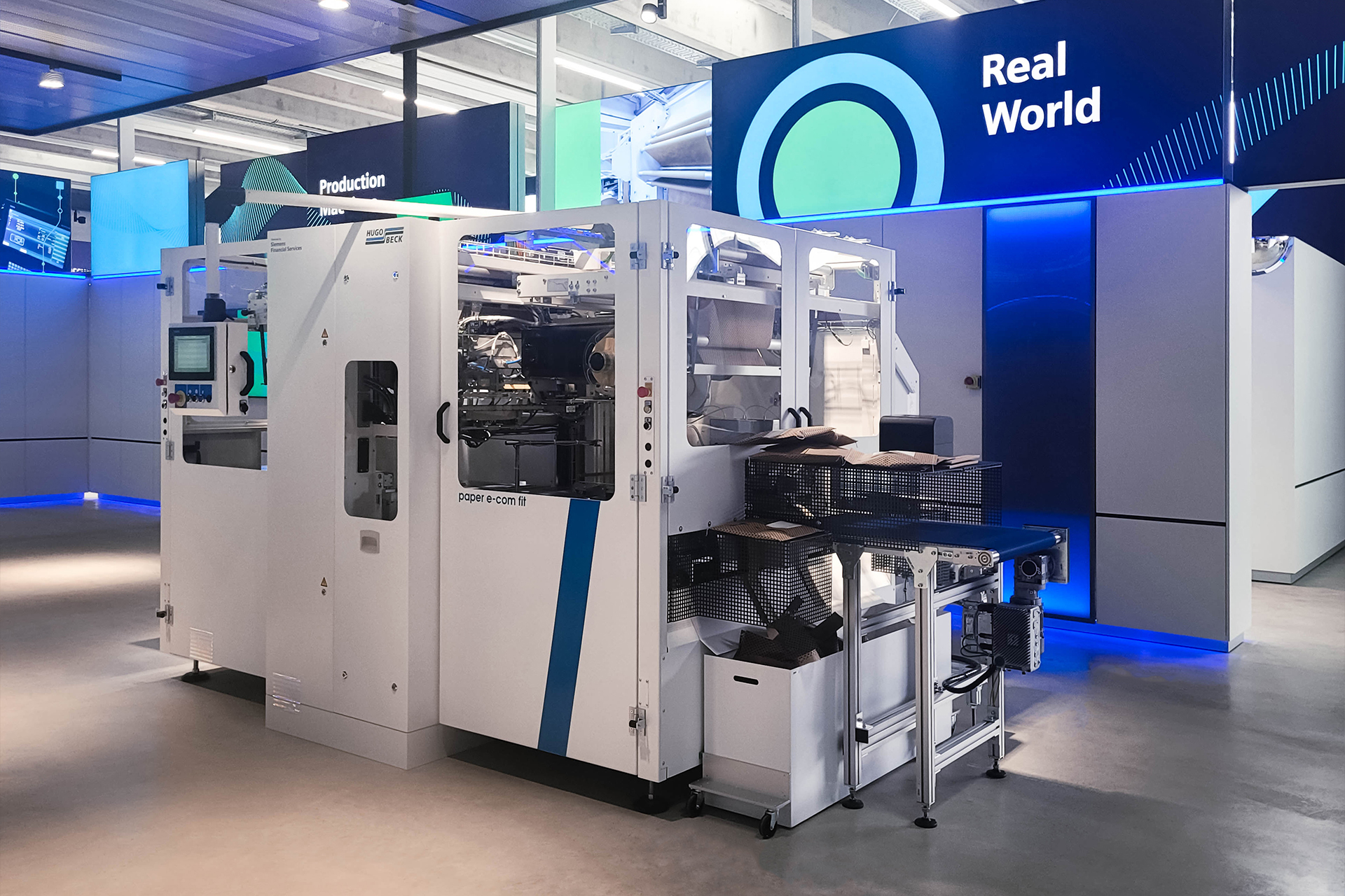 Industrielle Verpackungsmaschine im Showroom „Real World“, umgeben von Bildschirmen und LED-Wänden. Die Maschine verarbeitet Papierverpackungen und ist von modernen Präsentationselementen umgeben, die Digitalisierung und Produktion verbinden.