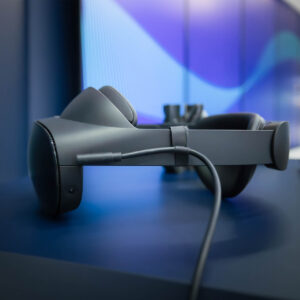 Seitenansicht einer modernen Virtual-Reality-Brille in dunklem Grau, liegend auf einer blauen Oberfläche vor einem abstrakten Hintergrund mit blauer Wellen-Grafik. Ein Anschlusskabel ist sichtbar mit dem Gerät verbunden. Die Szene vermittelt ein Hightech-Umfeld, vermutlich in einem Ausstellungs- oder Showroom-Kontext.