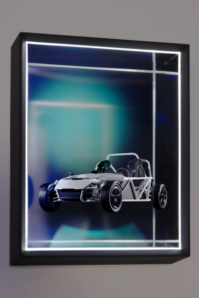 Holografisch wirkende 3D-Darstellung eines weißen Sportwagen-Modells (eRod) in einem beleuchteten, schwarzen Schaukasten mit Glasfront. Der Hintergrund ist weich verlaufend in Blau- und Grüntönen gehalten. Die Box wirkt modern, fast futuristisch, und betont die digitale Inszenierung des Fahrzeugs.