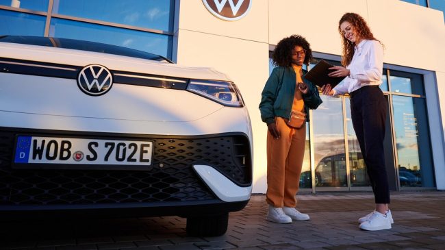 OneHub VW Servicemitarbeiterin zeigt der Kundin etwas auf dem Tablet, sie stehen neben einem VW Auto